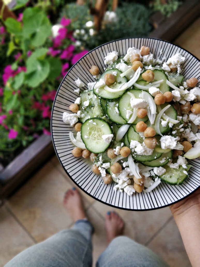 ein Teller mit Kichererbsensalat wird in der Hand gehalten, am Boden sind Füße zu sehen.